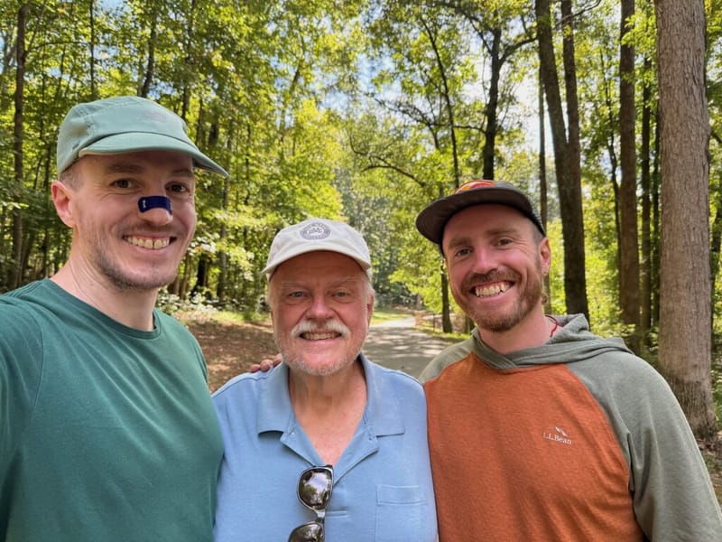 Three men smiling together outdoors in a wooded area