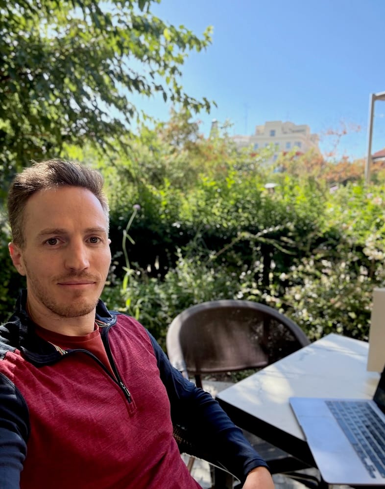Taylor with his laptop at a cafe table with nature in the background